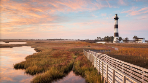 A scenic view of a lighthouse beside a marsh, with a wooden walkway and colorful sunset sky.