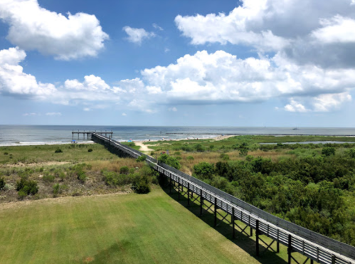 A wooden pier extends over a sandy beach, with lush greenery and a blue sky filled with clouds in the background.