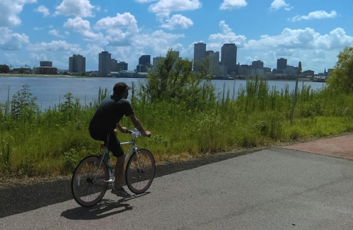 A person rides a bicycle along a path with a city skyline and river in the background under a blue sky with clouds.