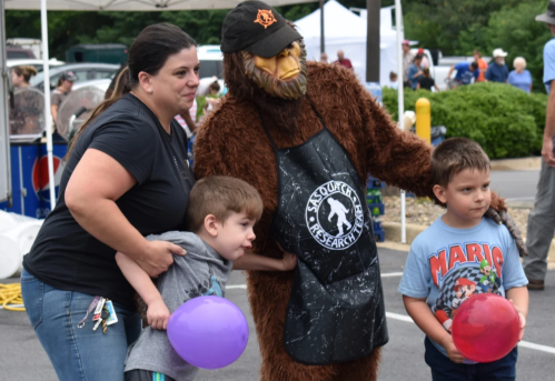 A woman and two children pose with a Bigfoot mascot at an outdoor event, balloons in hand, smiling for the camera.
