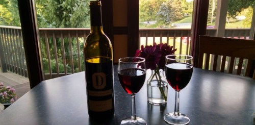A bottle of red wine with two glasses filled, set on a table with a vase of flowers and a scenic view outside.