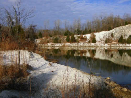 A serene winter landscape featuring a calm lake, snow-covered banks, and bare trees under a partly cloudy sky.