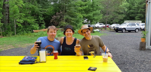 Three friends sit at a yellow picnic table outdoors, enjoying drinks in a wooded area.