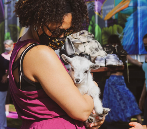 A person in a mask holds a small white goat, surrounded by colorful murals and people in the background.