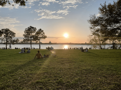 A sunset over a river with people sitting on the grass, enjoying the view and the warm evening light.
