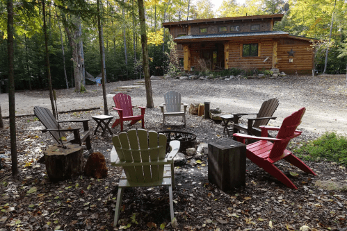 A cozy outdoor seating area with colorful chairs around a fire pit, surrounded by trees and a log cabin in the background.