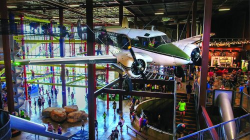 A vintage airplane hangs from the ceiling in a busy indoor space with people and play structures below.