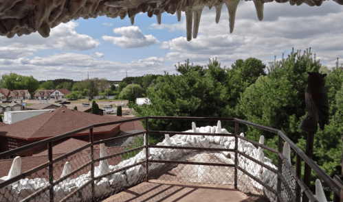 View from a lookout with a railing, featuring trees and buildings under a cloudy sky. Stalactites hang above.