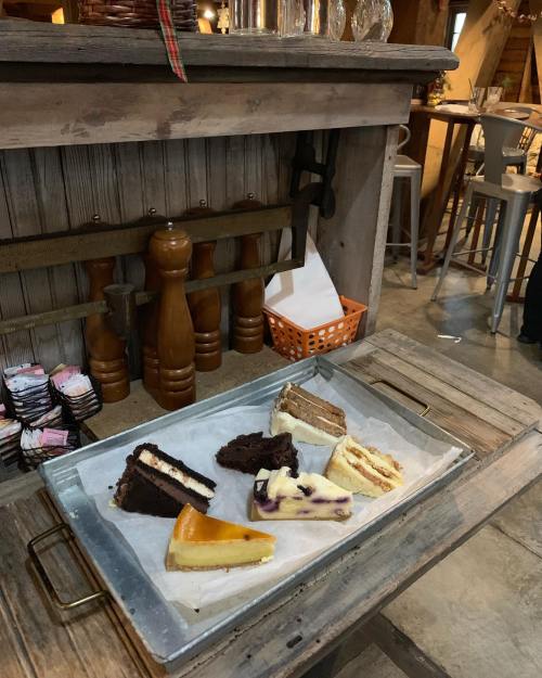 A wooden table displays an assortment of cakes on a tray, including chocolate, cheesecake, and layered desserts.