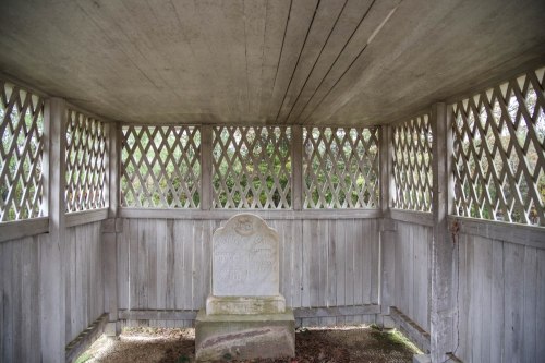 A weathered gravestone inside a wooden shelter with lattice walls, surrounded by greenery.
