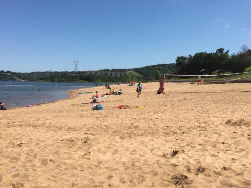 A sunny beach scene with people enjoying the sand and water, volleyball net in the background, and green hills nearby.