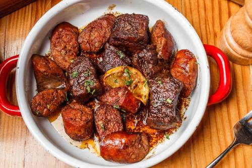 A white dish filled with various pieces of grilled meat, garnished with herbs and lemon slices, on a wooden table.