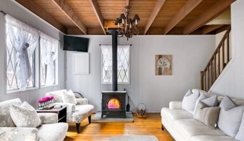 Cozy living room with a wood stove, comfortable seating, and large windows, featuring wooden beams and a warm atmosphere.