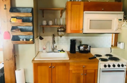 A cozy kitchen featuring wooden cabinets, a sink, coffee maker, microwave, and stove, with a wall-mounted storage rack.