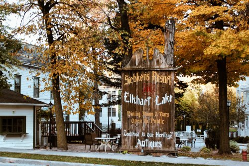 A rustic sign for Elkhart Lake's resort, surrounded by autumn foliage, highlighting dining and antique offerings.