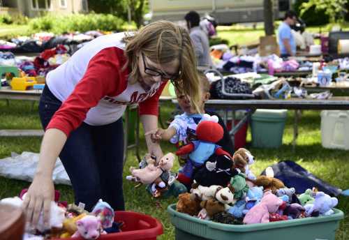 A woman sorts through a bin of stuffed animals while a child watches, surrounded by tables of toys in a sunny outdoor setting.