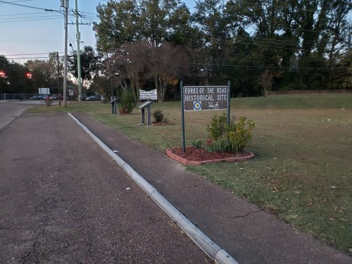 Sign for the Forks of the Road Historical Site, surrounded by grass and trees, with a sidewalk in the foreground.