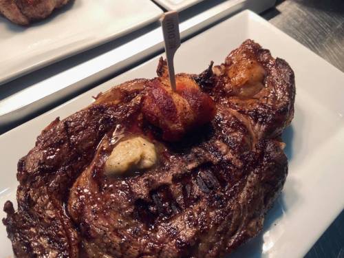 A juicy, grilled steak topped with a dollop of butter and a piece of bacon, served on a white plate.