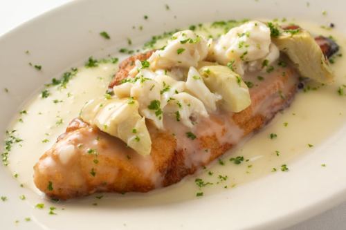 A plated fish fillet topped with crab meat and artichoke, drizzled with a creamy sauce and garnished with parsley.