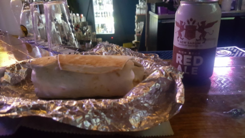 A burrito wrapped in foil next to a can of red ale on a bar counter.