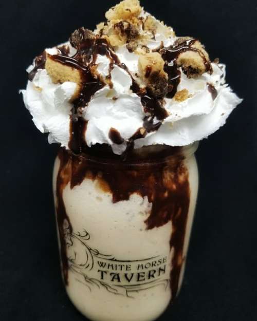 A creamy milkshake topped with whipped cream, chocolate drizzle, and cookie crumbles in a jar with a logo.