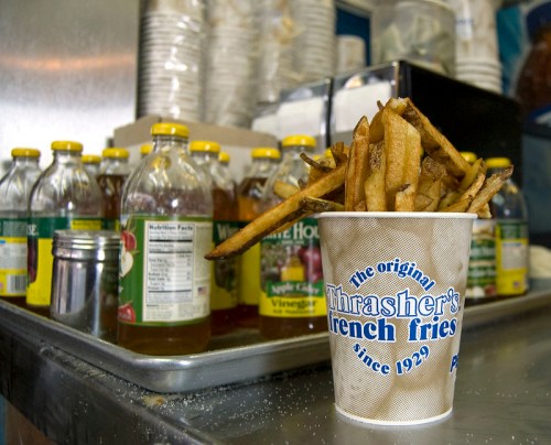A cup of golden French fries sits on a counter with bottles of vinegar in the background.