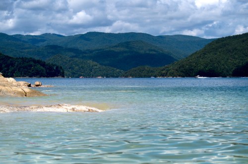A serene lake surrounded by mountains, with clear water and a rocky shoreline under a partly cloudy sky.