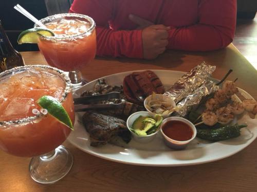 A platter of grilled meats, shrimp skewers, and sides, accompanied by two large cocktails with lime wedges.