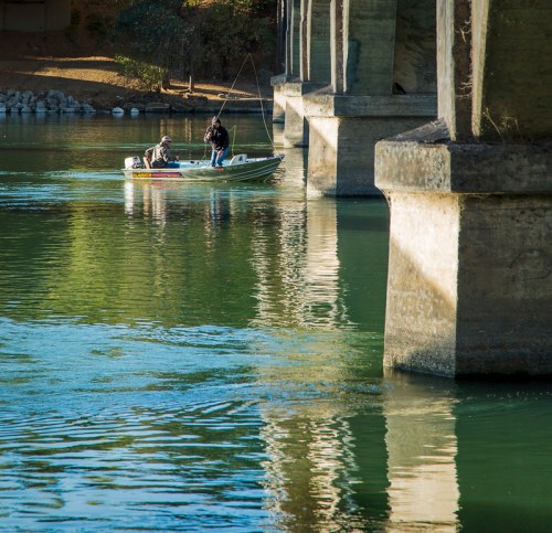 Two fishermen in a small boat under a bridge, casting lines into the calm water below. Reflections visible on the surface.