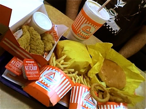 A tray of fast food featuring chicken strips, fries, a burger, onion rings, ketchup, and a drink.