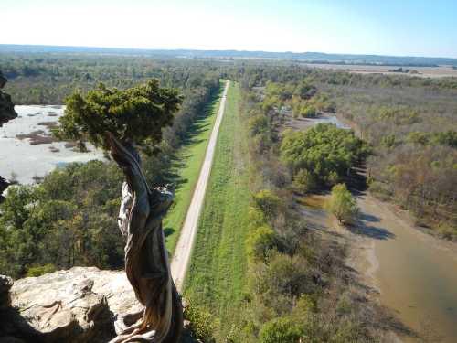 Aerial view of a winding dirt road bordered by lush greenery and a river, seen from a rocky overlook.