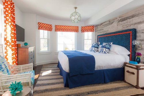 Bright bedroom with a blue bedspread, patterned curtains, and coastal decor, featuring a cozy seating area and wooden accents.