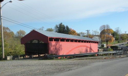 A red covered bridge with a metal roof, surrounded by trees and a rural landscape under a partly cloudy sky.