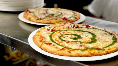 Two pizzas on white plates, one with a spiral of green sauce and cheese, on a stainless steel countertop.