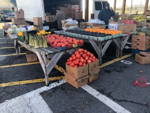 A farmers market with wooden tables displaying various fruits and vegetables, including tomatoes, oranges, and pineapples.