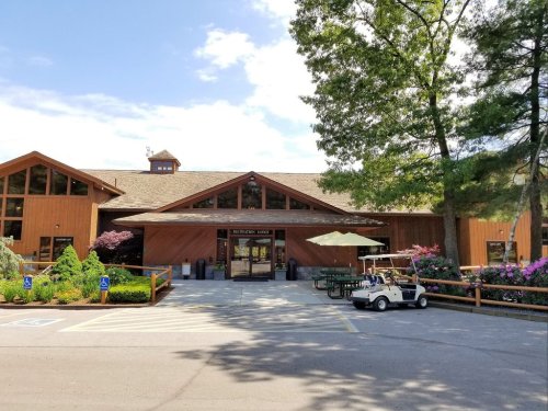 A wooden lodge with large windows, surrounded by greenery and flowers, featuring outdoor seating and a golf cart parked nearby.