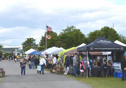A bustling outdoor market with vendors under tents, shoppers browsing, and trees in the background against a cloudy sky.