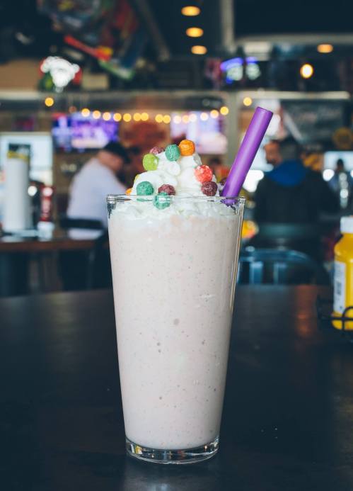 A tall glass of pink milkshake topped with whipped cream and colorful candy, served with a purple straw.