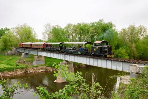 A vintage train crosses a bridge over a river, surrounded by lush greenery and trees on a cloudy day.