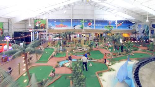 Indoor amusement park featuring mini-golf, palm trees, and colorful decorations, with families enjoying the space.