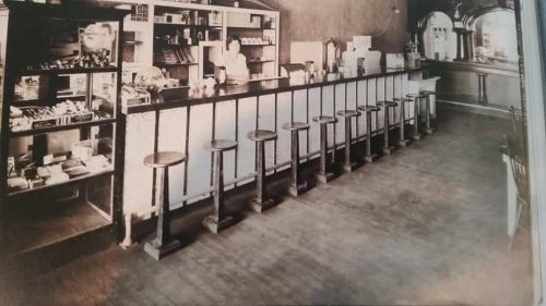 A vintage black-and-white photo of a diner counter with stools and shelves filled with products in the background.