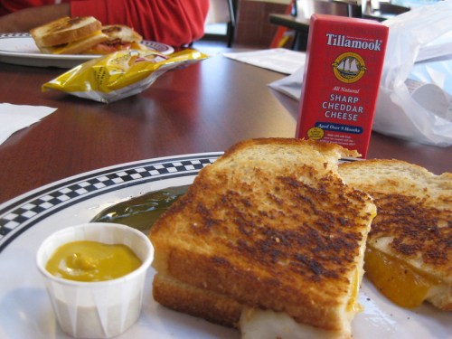 A plate with a grilled cheese sandwich, mustard, and a box of Tillamook sharp cheddar cheese on a table.