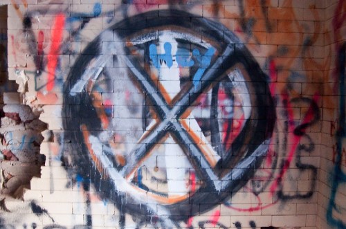 Graffiti of a large "X" symbol on a brick wall, surrounded by colorful paint splashes and markings.