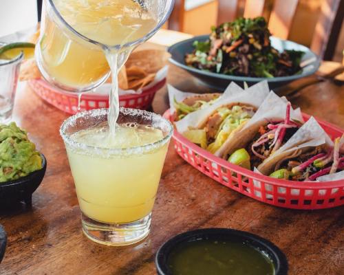 A pitcher pours a margarita into a glass with salt, surrounded by tacos, guacamole, and a salad on a wooden table.