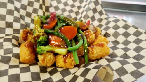 A serving of tater tots topped with mixed vegetables and a savory sauce, served in a checkered paper basket.