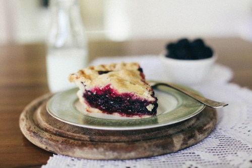 A slice of berry pie on a plate, with a glass of milk and a bowl of berries in the background on a wooden table.