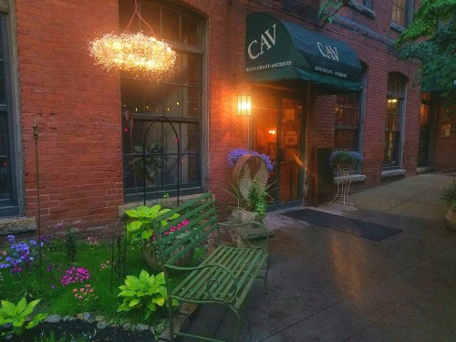 Charming restaurant entrance with a green bench, flowers, and a glowing chandelier in a brick alleyway.