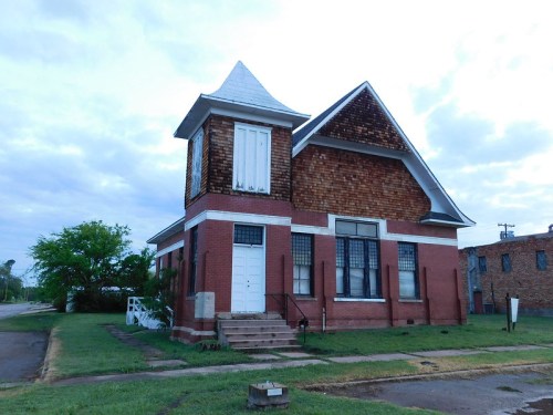 A two-story brick house with a unique peaked roof and wooden shingles, set on a grassy lot under a cloudy sky.