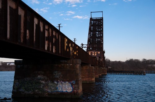 Rusty bridge structure over water, featuring graffiti on its supports and a clear blue sky in the background.