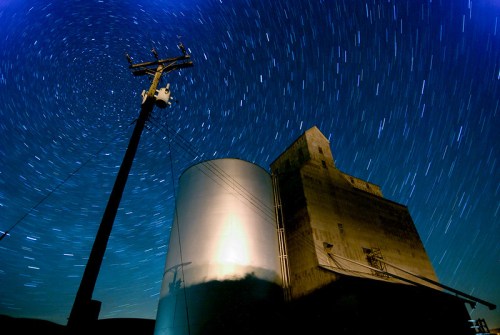 A grain silo under a starry night sky, with star trails creating circular patterns around a power pole.